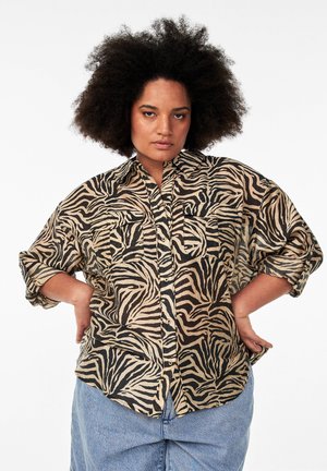 Chemise imprimée zèbre en beige et noir, avec une fermeture à boutons sur le devant, deux poches poitrine et des manches retroussées. Tissu doux et léger.