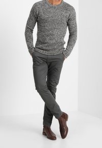 Šedý pletený svetr s kulatým výstřihem, kombinovaný s tmavě šedými kalhotami zúženého střihu a hnědými koženými kotníkovými botami. Viditelný detail texturované látky.