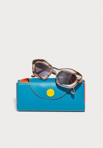 Occhiali da sole oversized con una fantasia a tartaruga sono posizionati sopra un brillante astuccio in pelle blu con dettagli arancioni e un logo circolare giallo.