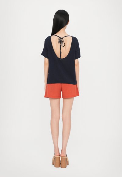 T-shirt navy con profonda scollatura sulla schiena, legato al collo, abbinato a pantaloni corti rossi. Il modello indossa scarpe con tacco a blocco beige. Tessuto morbido, design casual.