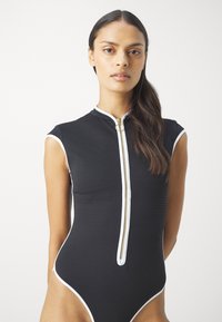 Schwarzer, gerippter Body mit mock neck, kurzen Ärmeln und einem goldenen Reißverschluss. Weiße Akzente umranden die Ärmel und Beinschnitte.