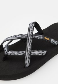 Teva sandals with black foam base and crisscross black and grey patterned straps. Features a small orange logo tag on the strap.
