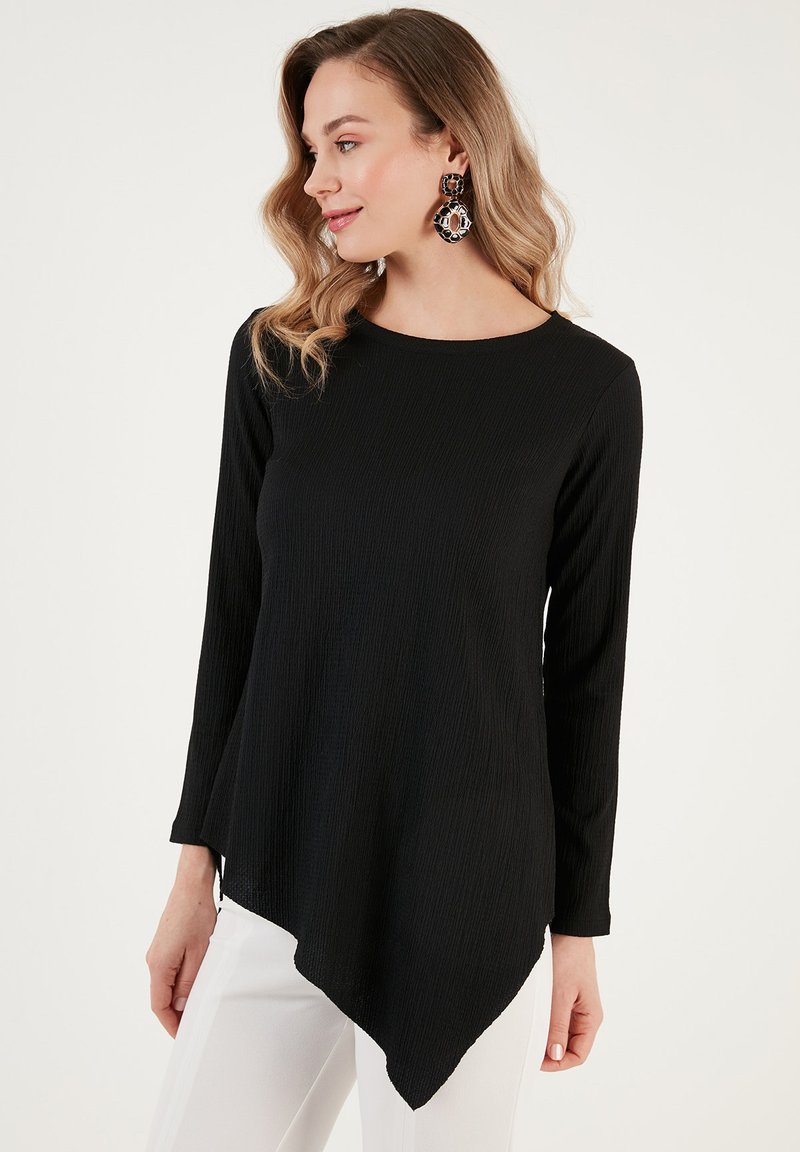 Top lungo nero a maniche con finitura testurizzata, caratterizzato da un orlo asimmetrico e un collo rotondo. Indossato con pantaloni bianchi.