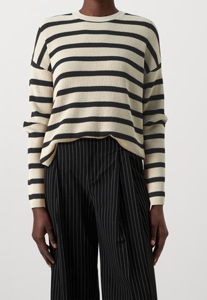 Persona con un jersey de rayas horizontales en crema y negro y pantalones negros con rayas verticales blancas, de pie frente a un fondo liso.