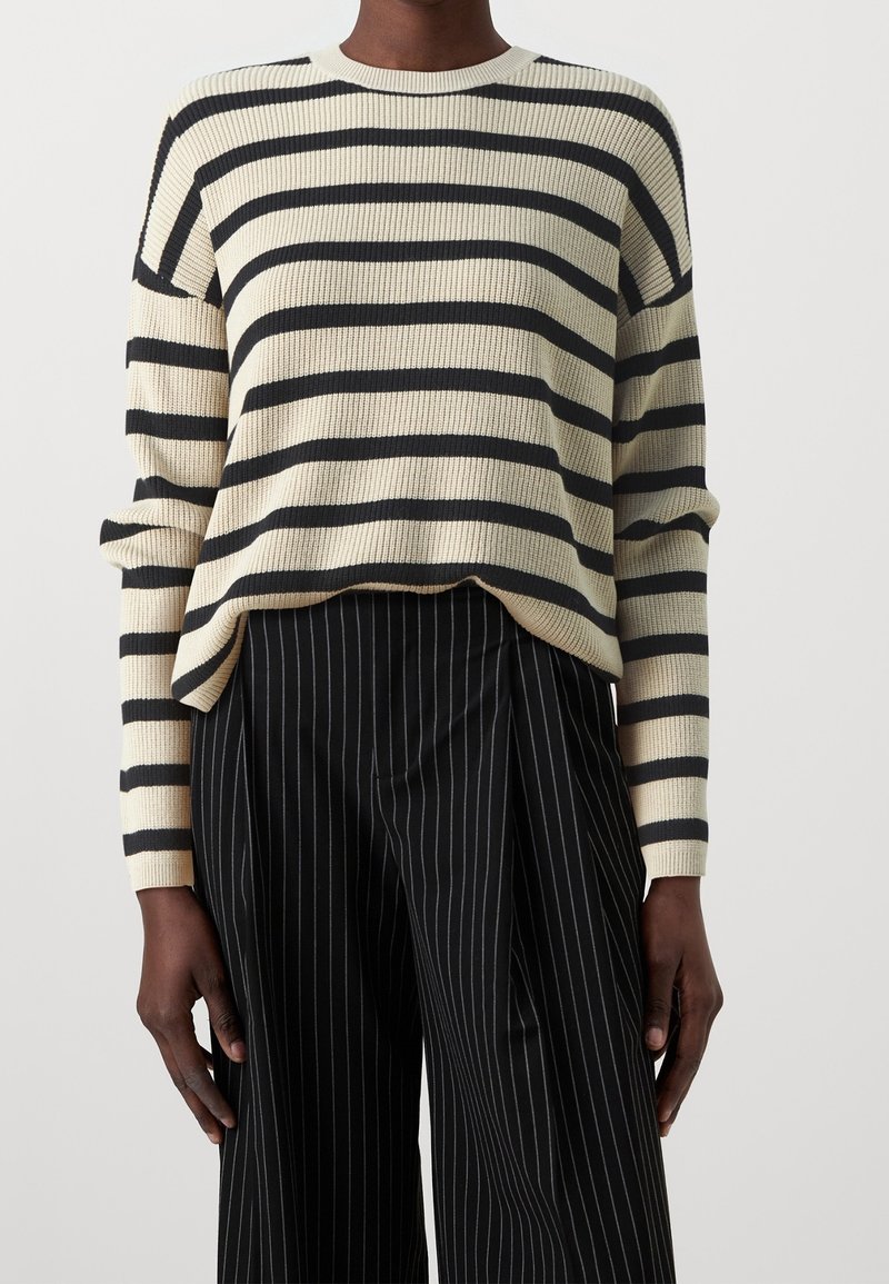 Personne portant un pull crème et noir à rayures horizontales et un pantalon noir avec des fines rayures verticales blanches, debout devant un fond uni.