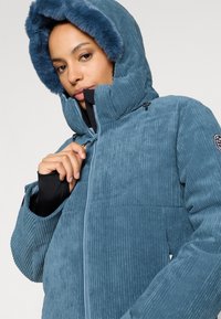 Modrá kordové sako s kapucí lemovanou umělou kožešinou, zipovým zapínáním a širokými rukávy s ribkovanou texturou a akcentem v podobě loga.