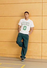 Hvid t-shirt med "CELTICS" og nummer "0" i grøn, sorte bukser, sorte sneakers, stående mod en træpanelvæg.