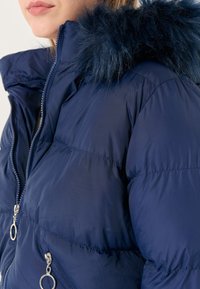 Giubbotto in nylon blu navy con cappuccio in pelliccia sintetica, chiusure con zip e texture trapuntata. Presenta due tasche frontali con zip e dettagli in metallo circolare.