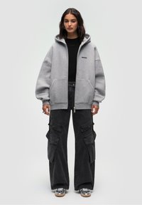 Felpa grigia oversize con zip, tasche frontali e logo ricamato; abbinata a pantaloni cargo neri larghi con multiple tasche.