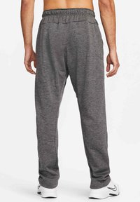Homme portant un pantalon de survêtement gris foncé et des baskets blanches Nike, debout les bras détendus, dos face à la caméra sur un fond blanc.
