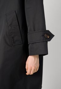 Black waterproof coat featuring a front pocket and buttoned cuff detail. Smooth texture with tailored fit and subtle branding.