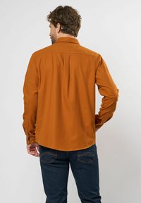 Bronzen blouse met lange mouwen en kraag, met knoopmanchetten en een rugpaneel detail. Draagt bij donkerblauwe jeans, met een gladde textuur.
