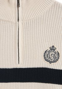 Stickad tröja i beige med en kvartdragkedja. Har en marinblå horisontell rand och en broderad logga på bröstet. Texturerad tyg.