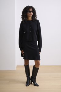 Černý pletený cardigan se zapínáním na knoflíky, v kombinaci s černou sukní ke kolenům. Modelka má na sobě černé vysoké kozačky na podpatku a sluneční brýle.