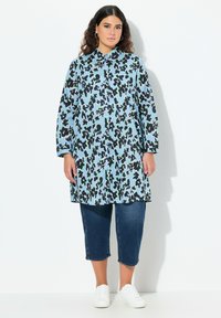 Camicia azzurra chiaro con un motivo astratto nero e verde, abbinata a pantaloni cropped in denim scuro e sneakers bianche.