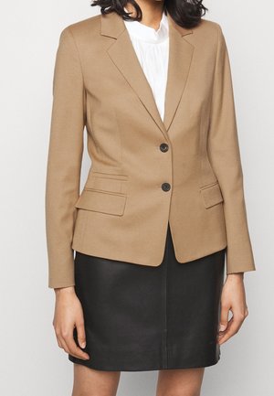 Tan blazer med en strukturerad passform, enknappsstängning, slagkrage och två framfickor. Bärs över en vit höghalsshals och en svart läderkjol.