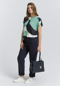 Vrouw staat in een groen, zwart en wit gestreept shirt, donkerblauwe broek, witte sneakers en houdt een zwarte handtas vast.