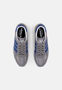 Pár šedých tenisek Adidas s modrými pruhy a tkaničkami, viděno shora na čistém bílém pozadí.
