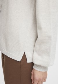 Ljust grått stickat tröja med ribbade mudd och nederkant, med en delad nederdesign och mjuk textur, parat med bruna tajta byxor.