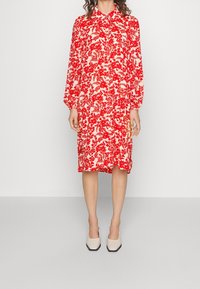  Robe imprimée rouge à fleurs avec des manches longues et un col, en tissu léger ; assortie à des chaussures à talons carrés beiges.