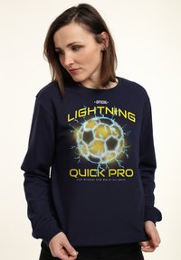 Frau mit schulterlangen braunen Haaren, die einen marineblauen Sweatshirt trägt, auf dem ein leuchtender Blitz-Fußball sowie der Text "Lightning Quick Pro" abgebildet sind.