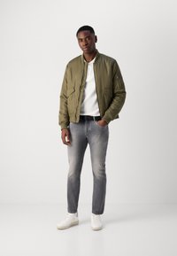 Jachetă bomber verde măsliniu cu fermoar, două buzunare frontale, purtată peste un tricou alb, combinată cu blugi gri și teniși albi.