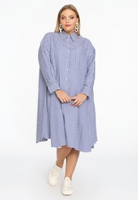 Gestreiftes Hemdkleid in Blau und Weiß mit übergroßer Passform, Knopfleiste, Kragen, langen Ärmeln und abgerundetem Saum. Getragen mit weißen Sneakern.