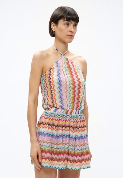 Γυναίκα με κοντά σκουρόχρωμα μαλλιά, φοράει αμάνικο τοπ με σχέδιο ζικ-ζακ σε πολλαπλά χρώματα και ταιριαστή φούστα ψηλόμεση, σε λευκό φόντο.