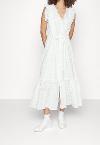 Vit bomullsklänning med broderade detaljer, volangärmar, midjeband och en flerlagerskjol; kombinerad med vita sneakers för en avslappnad look.