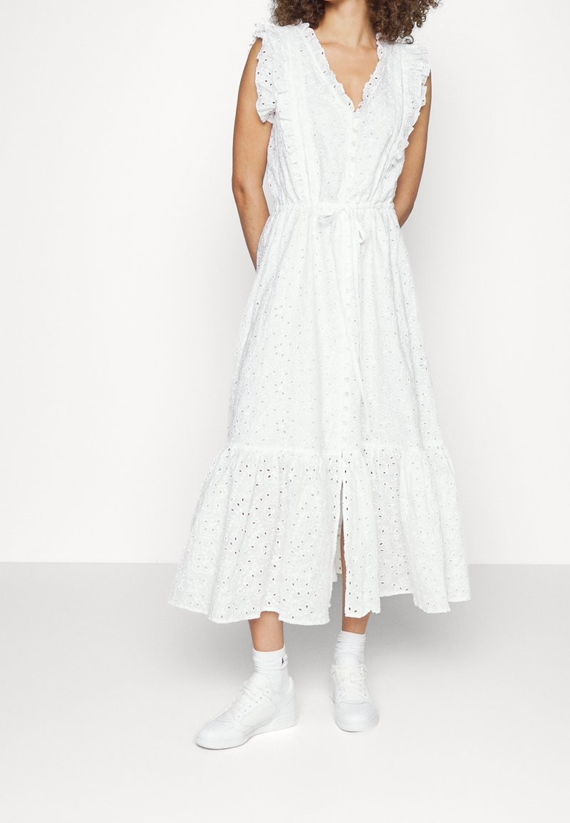 Vit bomullsklänning med broderade detaljer, volangärmar, midjeband och en flerlagerskjol; kombinerad med vita sneakers för en avslappnad look.