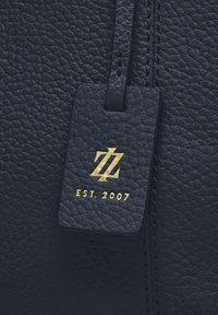 Close-up van zwart textuurleer met een gestikt label in goud dat een logo en de tekst "EST. 2007" toont.