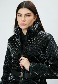 Chaqueta acolchada negra de material brillante con cuello alto, cierre de cremallera y detalle de cinturón en la cintura. Textura suave con patrón de diamantes.
