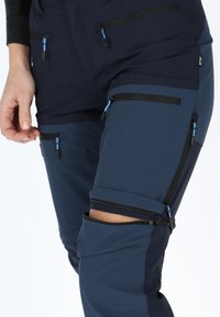Navy outdoor pants featuring multiple zippered pockets, contrasting fabric panels, smooth texture, and adjustable cuffs with zippers.