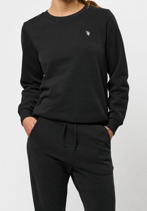 Femme portant un sweat-shirt noir à manches longues et un pantalon de jogging noir assorti, les mains dans les poches sur un fond uni.