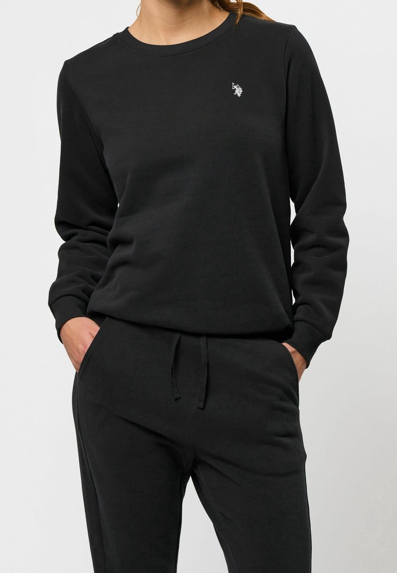 Femme portant un sweat-shirt noir à manches longues et un pantalon de jogging noir assorti, les mains dans les poches sur un fond uni.