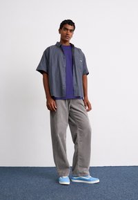 Camisa de manga curta, com botões, em padrão xadrez azul e verde, sobre uma t-shirt roxa. Calças de veludo cotelê cinzas e ténis azuis completam o conjunto.