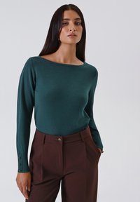 Jonge vrouw met lang donker haar, gekleed in een teal lange mouwen top en hoge taille bruine broeken, staande met één hand in de zak.