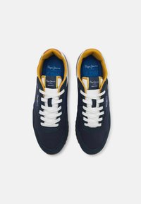 Sötétkék sneaker fehér fűzőkkel, sárga szegéllyel és barna gumitalppal. Szövet felsőrésszel rendelkezik, valamint logóval az oldalon és a nyelvén.