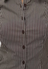 Ruskeanraitaista napillista paitaa, jossa on ohuita valkoisia pystysuoria viivoja, kaksi ruskeaa nappia ja sileä kangasnäkymä.