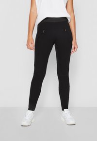 Leggings noirs au design ajusté, avec des poches zippées sur les côtés, des panneaux texturés et une large ceinture. Le tissu est lisse et extensible.