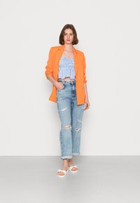 Blazer laranja, top curto floral azul claro, jeans rasgados azul claro e sandálias chunky brancas. Modelo posa com as mãos nos bolsos.