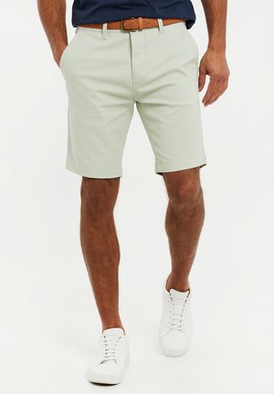 Pantalones cortos verdes claros con corte recto, bolsillos laterales y dobladillo doblado, usados con un cinturón trenzado marrón y zapatillas blancas.