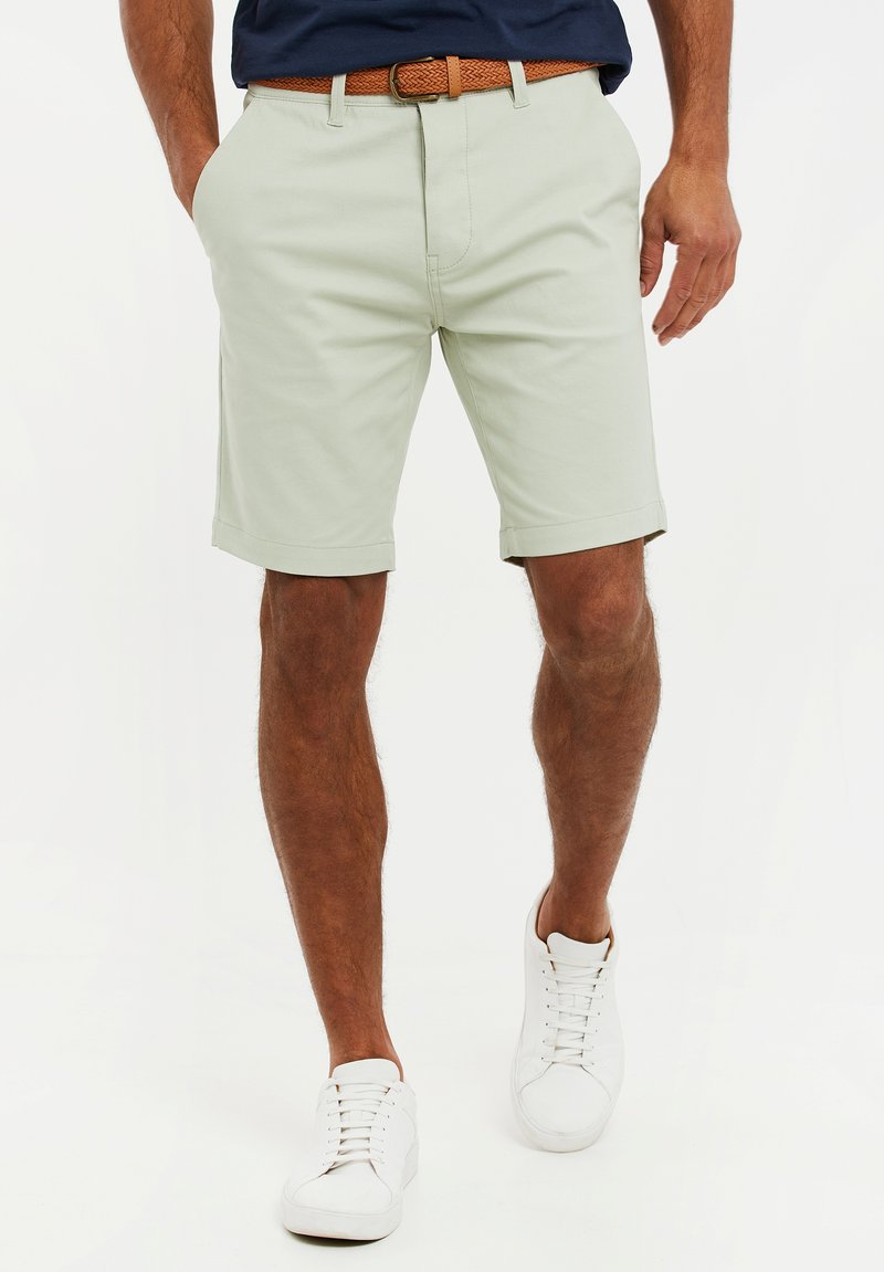 Pantalones cortos verdes claros con corte recto, bolsillos laterales y dobladillo doblado, usados con un cinturón trenzado marrón y zapatillas blancas.