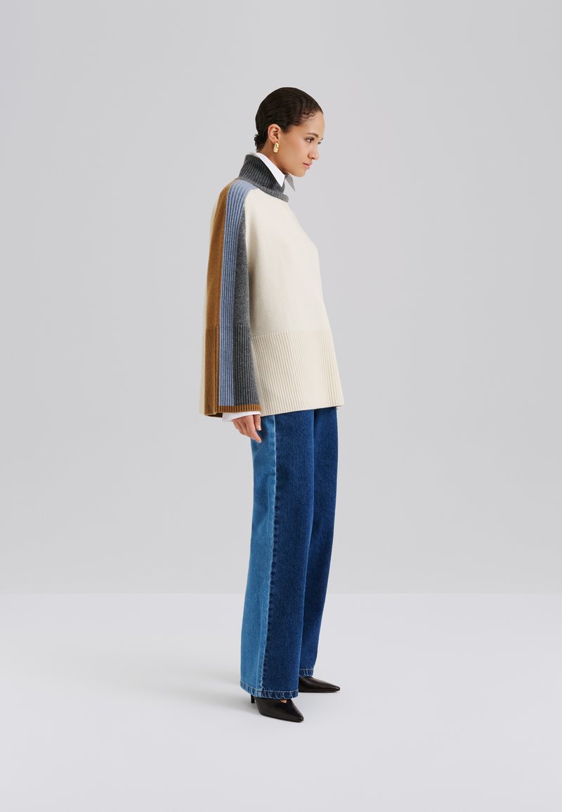Pull asymétrique en crème, bleu, gris et beige ; texture côtelée ; col haut ; associé à un jean bleu ample et des chaussures noires à bouts pointus.