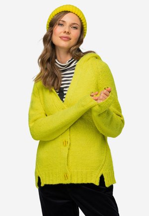 Cardigan lavorato a maglia verde lime con due bottoni gialli, scollo a V e fessure laterali. Abbinato a un dolcevita a righe bianche e nere sotto.