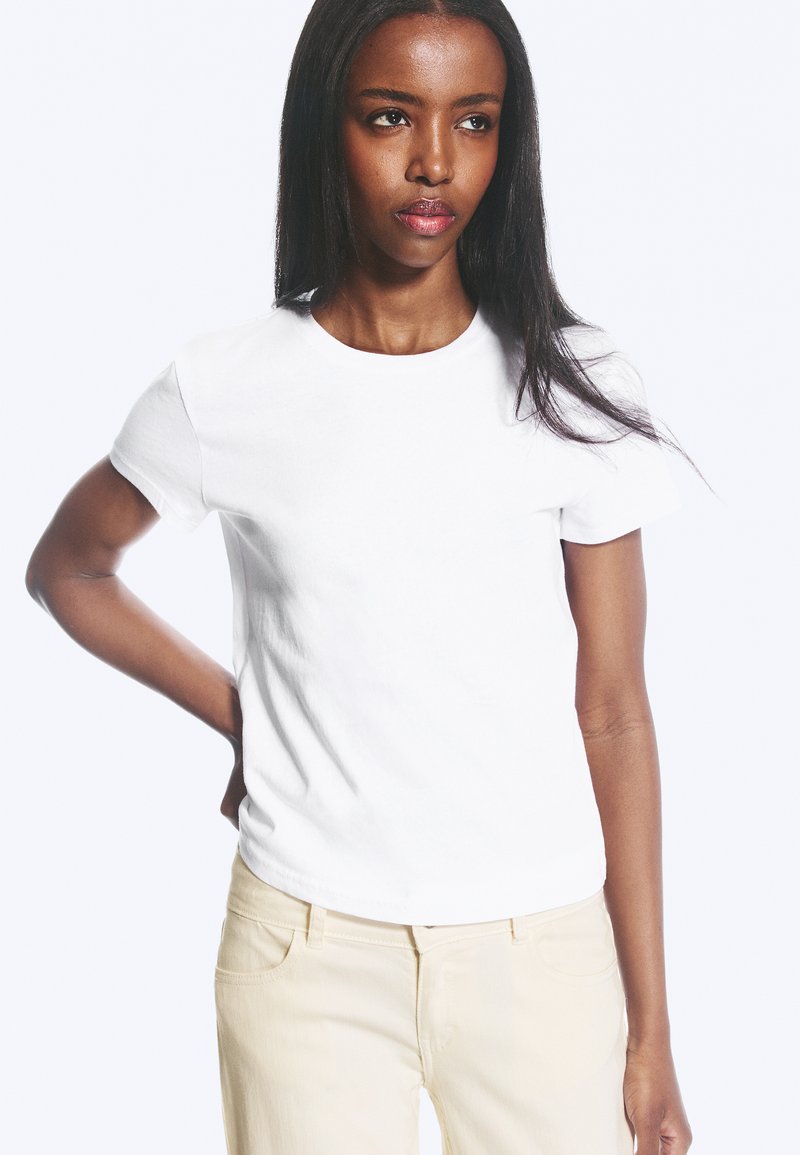 Femme aux longs cheveux foncés portant un t-shirt blanc uni à manches courtes et un pantalon beige clair, debout avec une main sur la hanche.