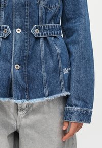 Τζιν μπουφάν με μεταλλικά κουμπιά πίεσης, ρυθμιζόμενες πλαϊνές λωρίδες, ξεφτισμένο τελείωμα και κέντημα "KARL LAGERFELD JEANS" στη δεξιά πλευρά.