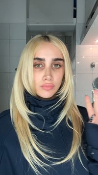 Jeune personne aux longs cheveux blonds et aux yeux verts portant une veste sombre, levant la main à l'intérieur, avec des murs carrelés en arrière-plan.