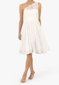 Bílé šaty s jedním krajkovým ramínkem, skládáním sukně a přiléhavým živůtkem. V kombinaci s bíle doplněnými sandály na podpatku, které zdůrazňují hladkou strukturu.