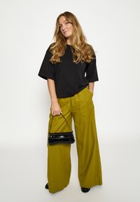 Must suur t-särk, rohelised laiade säärega püksid, särav must käekott, mustad kingad. Modell seisab neutraalsel taustal, käed taskutes.